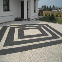 Ułożona kostka brukowa w geometryczny wzór przed domem z białymi ścianami i ciemną stolarką okienną i drzwiową. Ciemne pasy kostki tworzą kwadratowy obrys na jasnej nawierzchni, prowadzący do wejścia.