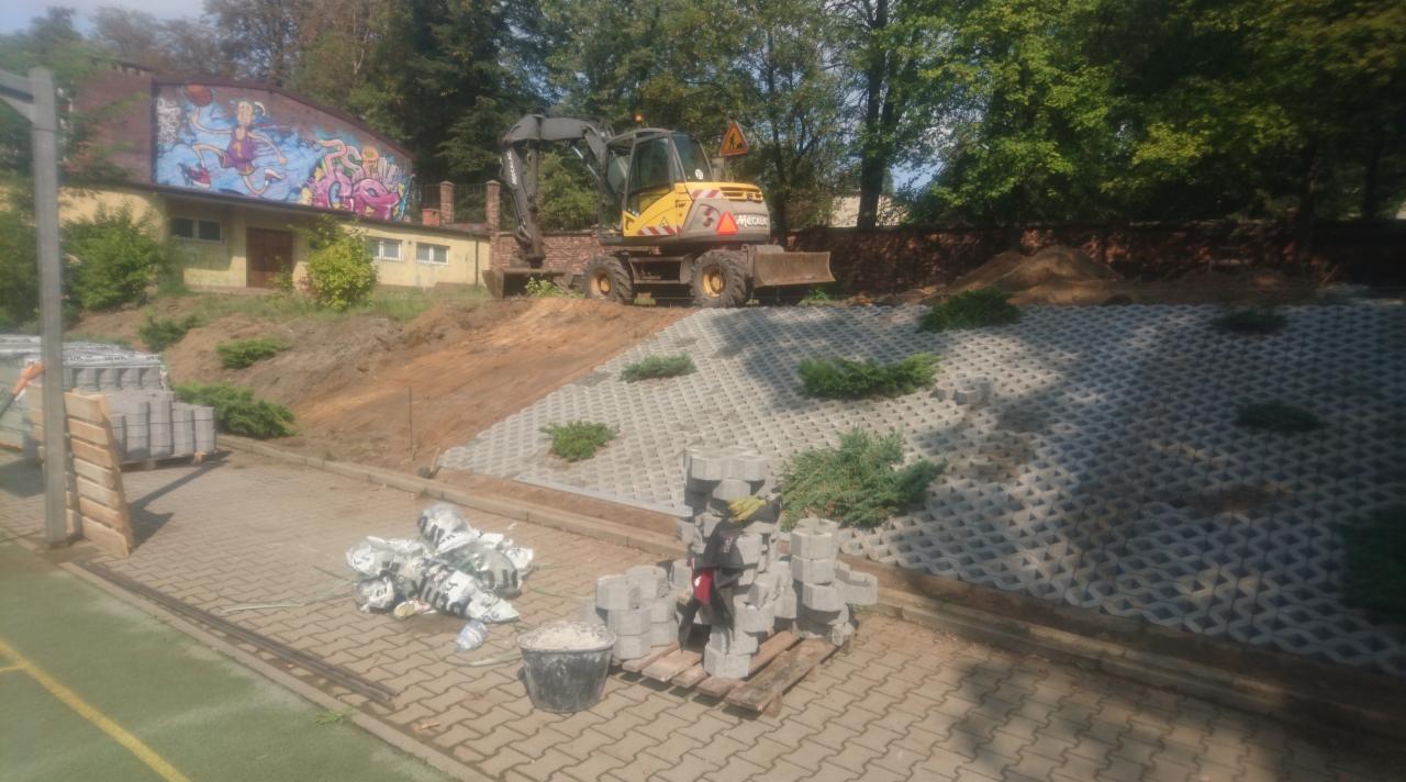 Żółta koparka Mecalac na wzgórzu z ułożoną ażurową kostką betonową, w tle budynek z muralem i sterty kostki brukowej na paletach.