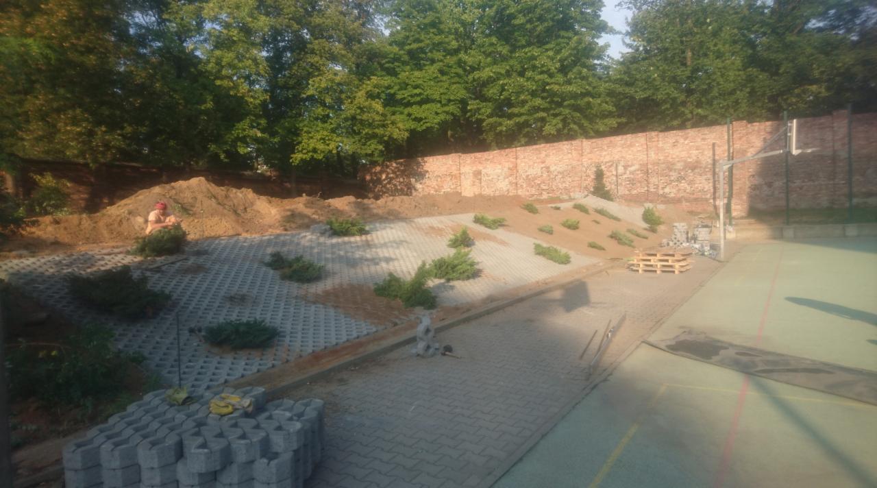Ukończony fragment parkingu z ażurowych płyt betonowych, obsadzony niskimi krzewami iglastymi na usypanym nasypie ziemnym, w tle ceglany mur i drzewa.