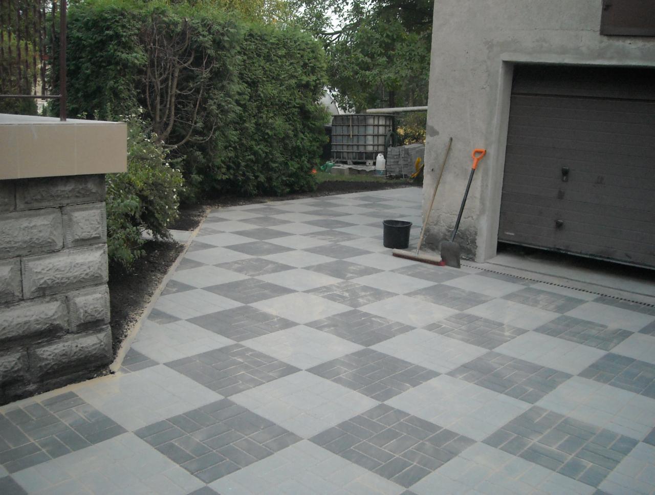 Ułożona nawierzchnia z kostki brukowej w szachownicę, widok na garaż, z boku mur z kamienia, w tle zieleń.