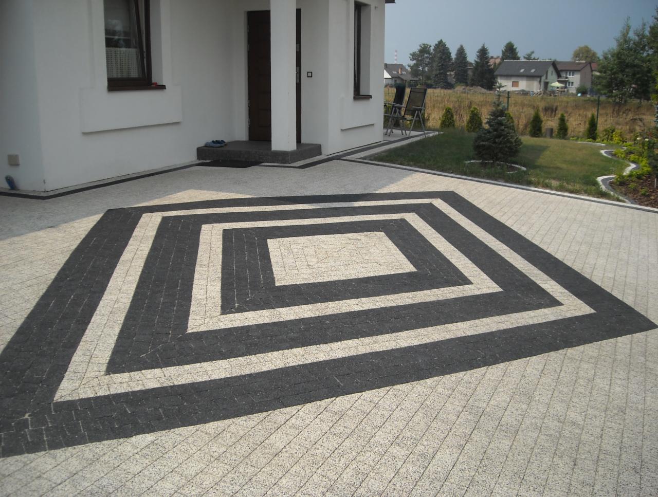 Ułożona kostka brukowa w geometryczny wzór przed domem z białymi ścianami i ciemną stolarką okienną i drzwiową. Ciemne pasy kostki tworzą kwadratowy obrys na jasnej nawierzchni, prowadzący do wejścia.