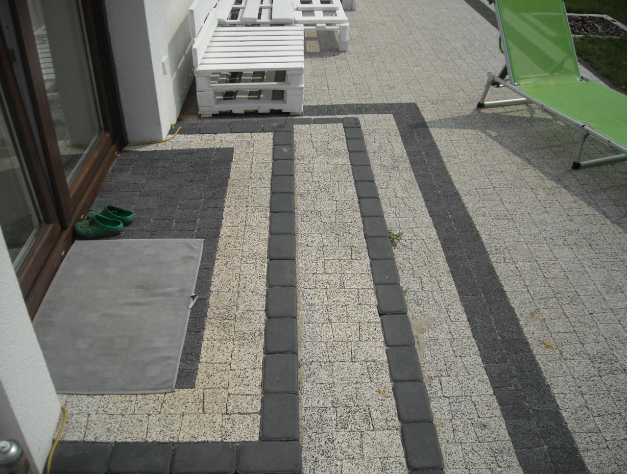 Ułożona kostka brukowa w odcieniach szarości i beżu tworząca wzór na podwórku, z widocznym fragmentem białych mebli ogrodowych i zielonym leżakiem w tle, oraz zielonymi butami przy wejściu do domu.