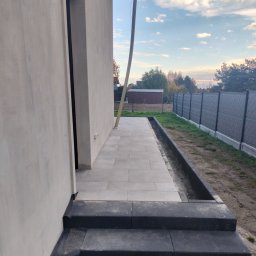Bruk-Tim - Jasnoszara kostka brukowa ułożona jako chodnik przy budynku z dwoma stopniami wejściowymi. Widoczne obrzeża i fragment ogrodzenia panelowego.