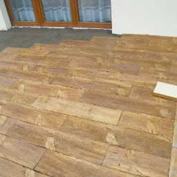 Fragment układanej powierzchni z kostki imitującej drewno, widoczny fragment ściany budynku i okno, w tle podsypka piaskowa.