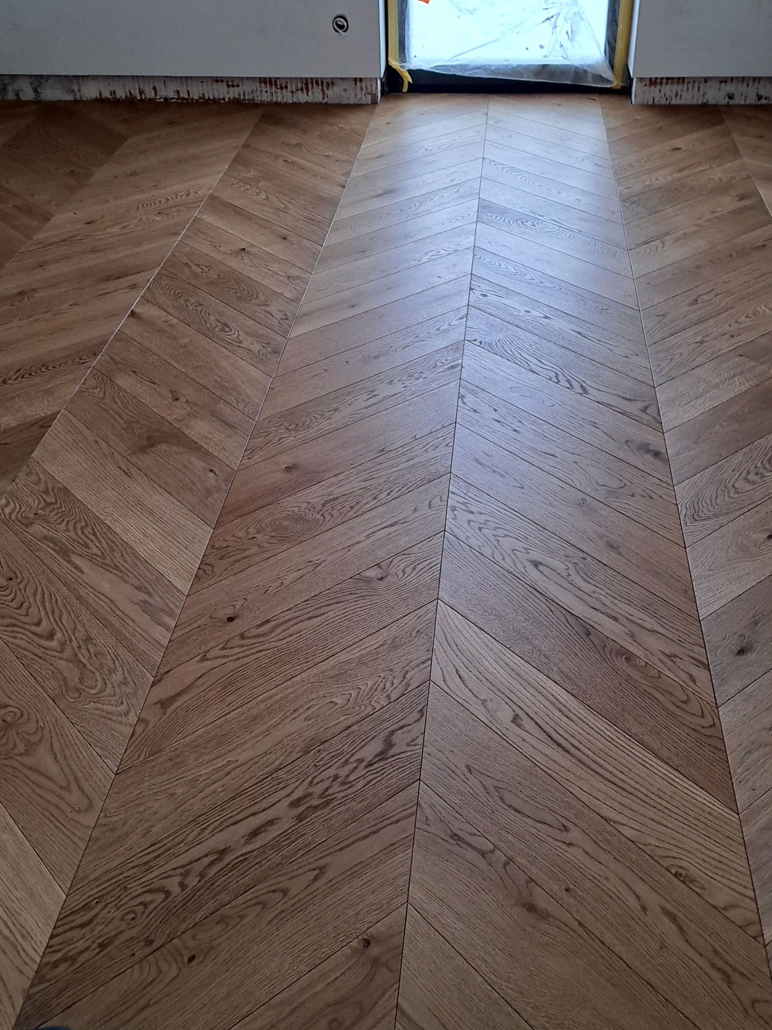 Ułożona podłoga z drewnianych klepek w jodełkę, widoczne zabezpieczone folią okno i fragment ściany z widocznymi śladami po pracach wykończeniowych.