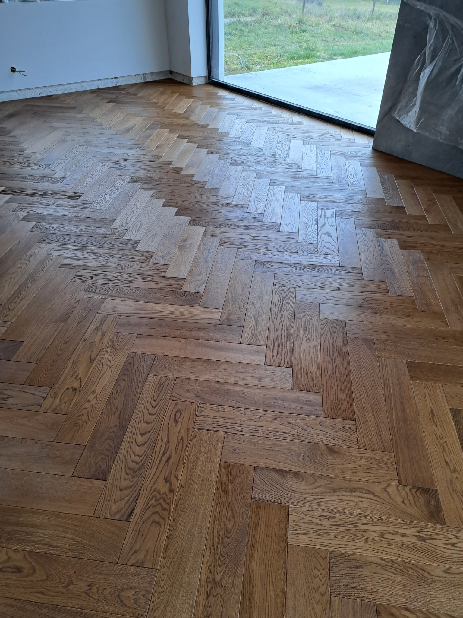 Ułożony parkiet w jodełkę francuską, widoczne słoje drewna, jasne ściany i duże okno w tle.