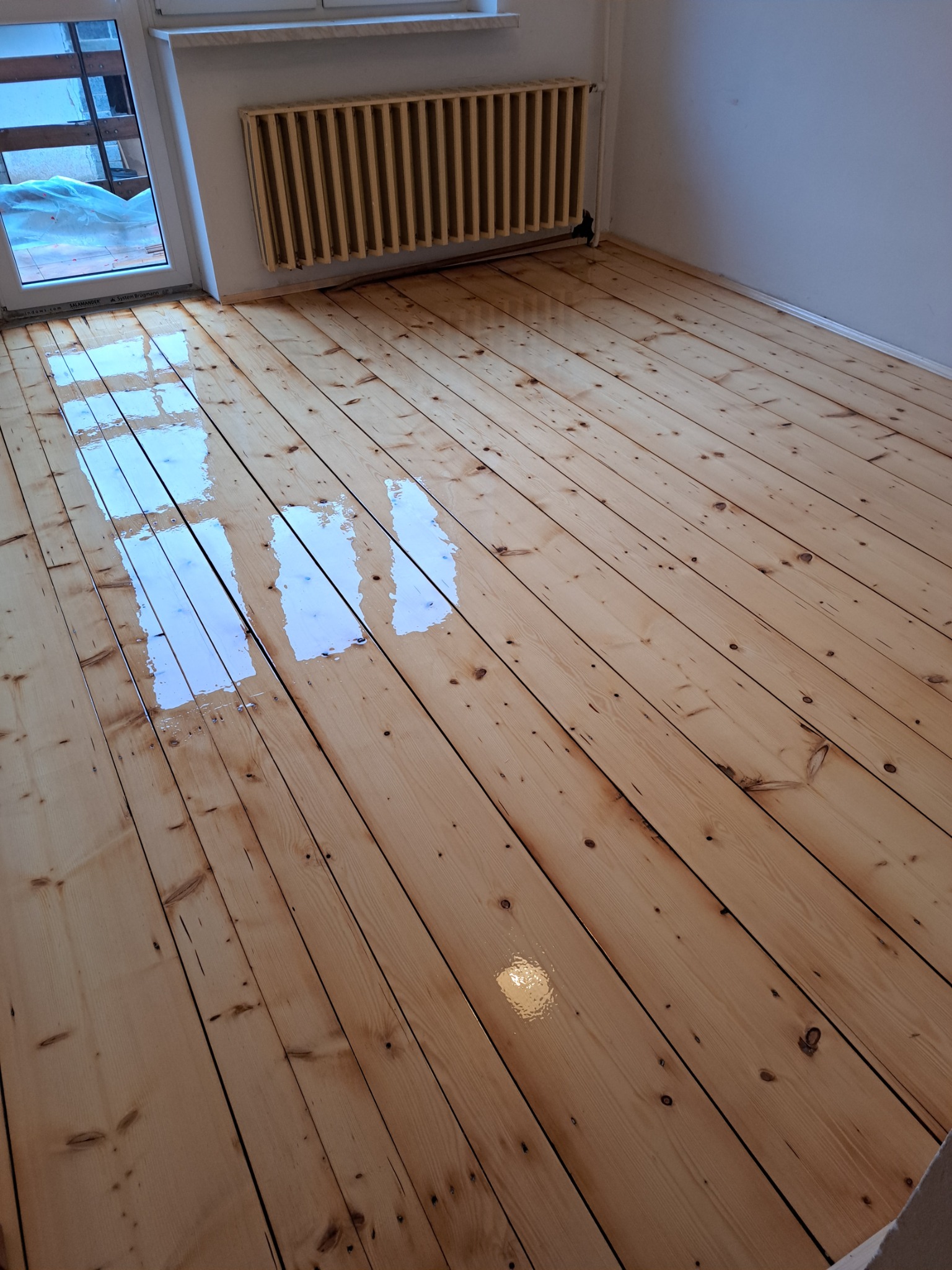 Świeżo polakierowana, jasna podłoga drewniana z widocznymi refleksami światła, grzejnik żeberkowy przy ścianie, okno balkonowe w tle.