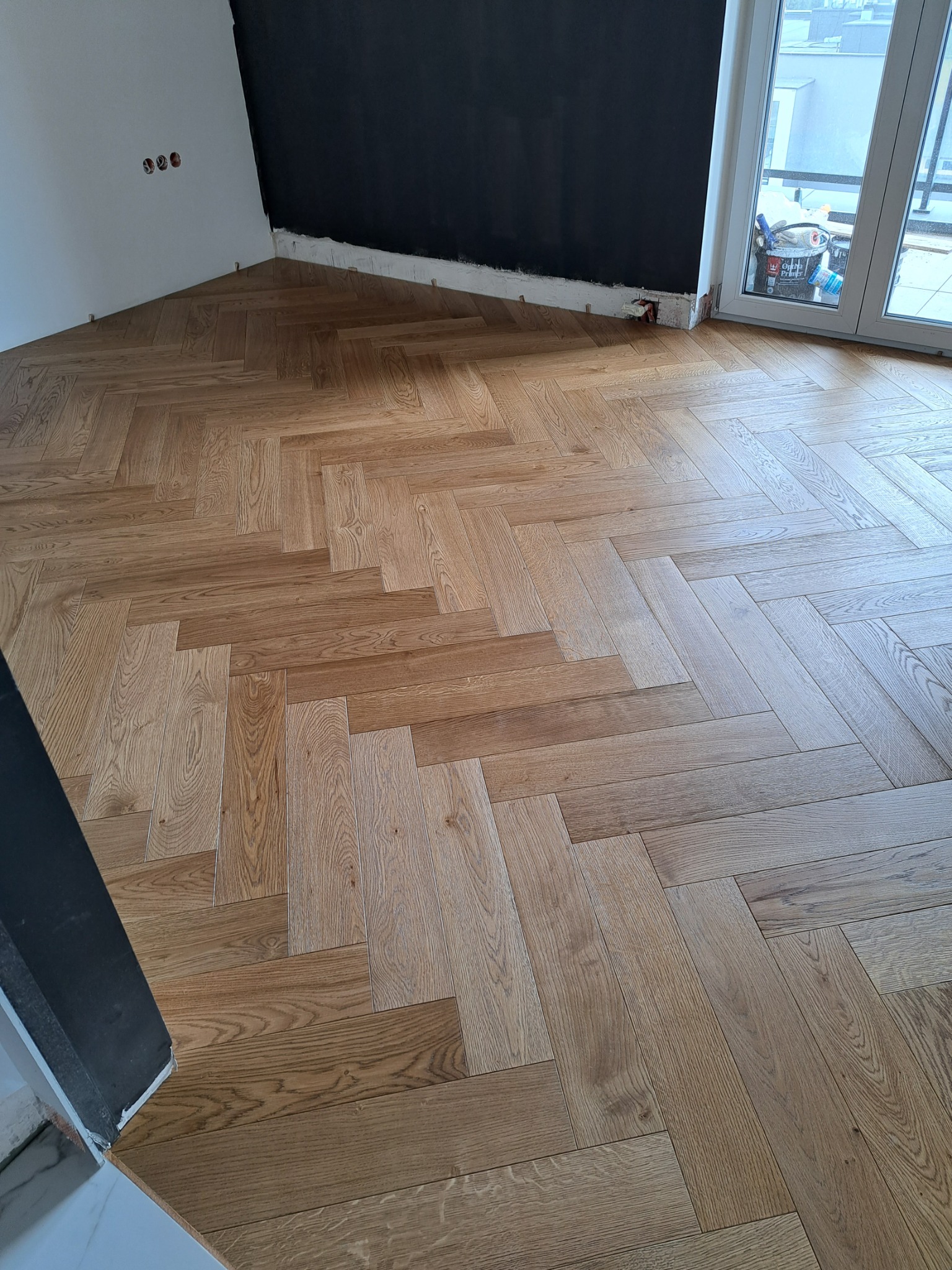 Ułożony parkiet w jodełkę w Wiśniowej, widoczne przygotowane ściany do malowania i drzwi balkonowe. Podłoga z jasnego drewna.