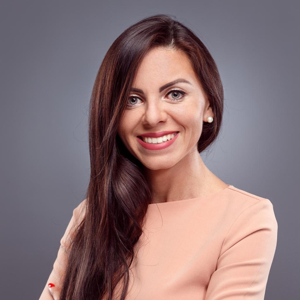 Portret uśmiechniętej kobiety z długimi, ciemnymi włosami, ubranej w pastelową sukienkę na tle szarego, rozmytym tle.