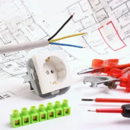 Białe gniazdko elektryczne, przewód z kolorowymi żyłami, zielona listwa zaciskowa oraz narzędzia na tle planu architektonicznego.