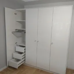 Biała szafa z frontami na wysoki połysk, częściowo otwarta, ukazująca wysuwane szuflady i p&oacute;łki z mechanizmem.