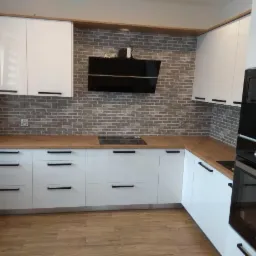 Biała zabudowa kuchenna z drewnianym blatem, czarnymi uchwytami i okapem na tle ściany z cegły. Widoczna płyta indukcyjna i piekarnik.