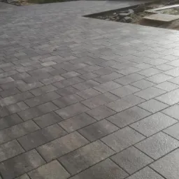 Ułożona szara kostka brukowa w jodełkę, z widocznymi fugami, fragmentem osoby po lewej stronie i przygotowanym terenem pod dalsze prace po prawej stronie.