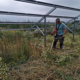 M&M Wycinka i pielęgnacja drzew - Mężczyzna w słuchawkach ochronnych kosi trawę i chwasty pod metalową konstrukcją, prawdopodobnie pod budowę paneli słonecznych. W tle widoczny krajobraz polny pod pochmurnym niebem.