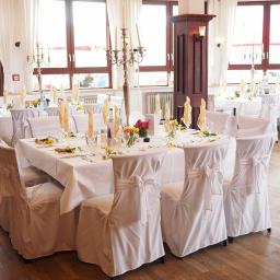 DogataStudio Agata Płocińska - Sala bankietowa przygotowana na uroczystość: białe obrusy, krzesła w pokrowcach z kokardami, wysokie świeczniki, kwiaty w wazonach i żółte serwetki na stołach, widok z parkietu.