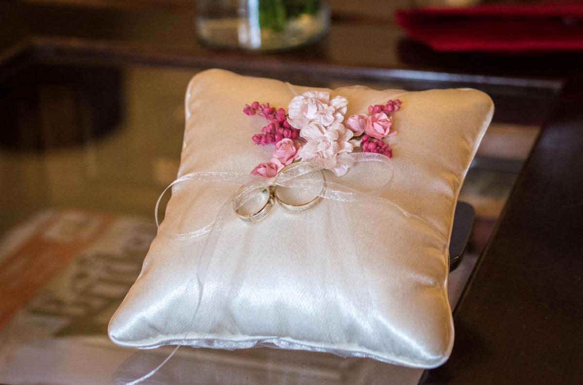Poduszka na obrączki z delikatnymi różowymi i białymi kwiatami, leżąca na szklanym stole w Warszawie. Obrączki zawiązane białą, cienką wstążką.