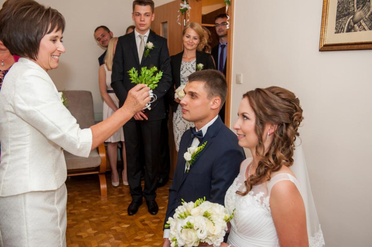 Błogosławieństwo Młodej Pary przez kobietę w białym kostiumie, w tle goście weselni obserwujący uroczystość, bukiet białych kwiatów w dłoniach nowożeńców.