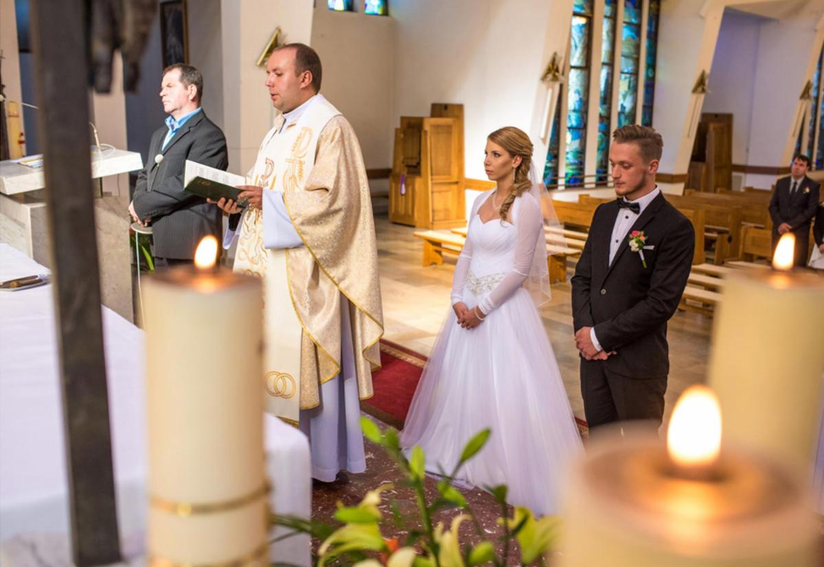 Para młoda podczas ceremonii ślubnej w kościele, ksiądz czyta fragment z księgi, w tle witraże i ławki.