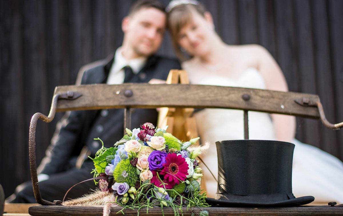 Para młoda w tle, na pierwszym planie bukiet ślubny z różowymi i fioletowymi kwiatami oraz czarny cylinder na drewnianej konstrukcji.