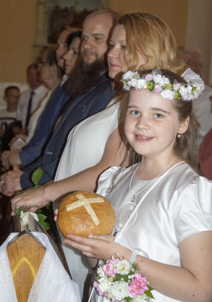 Dziewczynka w białej sukience komunijnej i wianku na głowie trzyma chleb z krzyżem, w tle rodzina podczas ceremonii w kościele.
