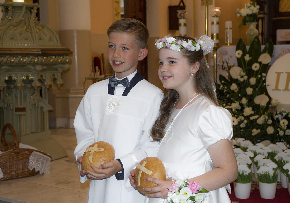 Dwoje dzieci, chłopiec w muszce i dziewczynka z wiankiem, w białych strojach komunijnych trzymają chleb z krzyżem w kościele.