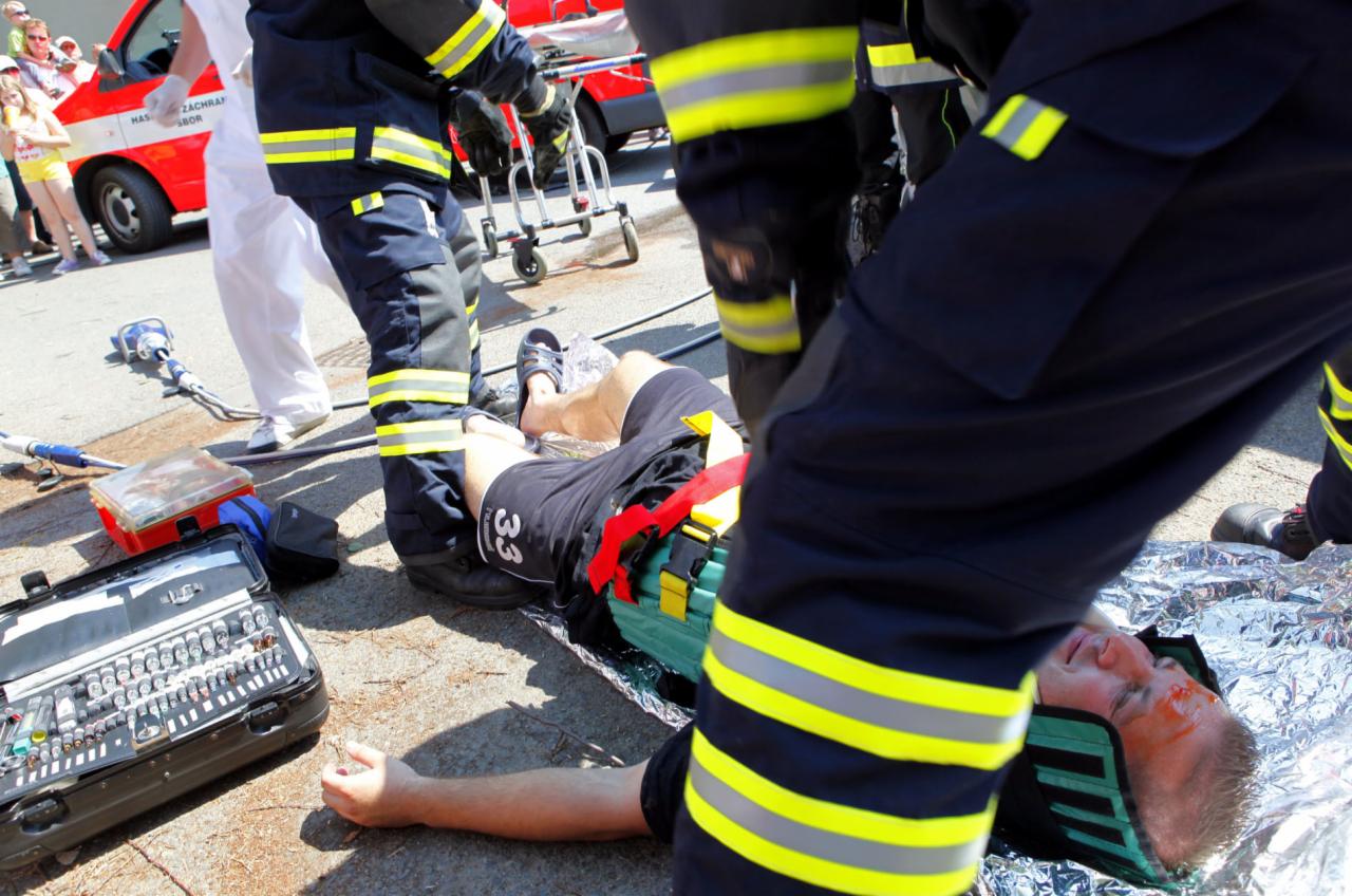 Ratownicy udzielają pomocy poszkodowanemu mężczyźnie leżącemu na kocu termicznym, widoczna otwarta apteczka i ambulans w tle.