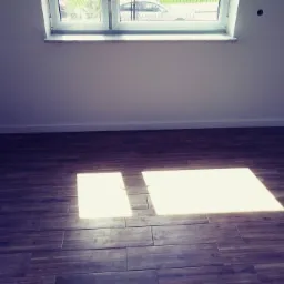 Ułożona podłoga z płytek imitujących drewno w pustym pokoju w Częstochowie, z oknem dachowym i plamami światła słonecznego.