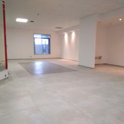 Remont-ownia - Przestronne, jasne wnętrze biurowe z beżową podłogą z płytek, białymi ścianami, kolumnami i oknem. W podłodze widoczny prostokątny obszar z innym odcieniem płytek. Na ścianie widoczne punkty świetlne.