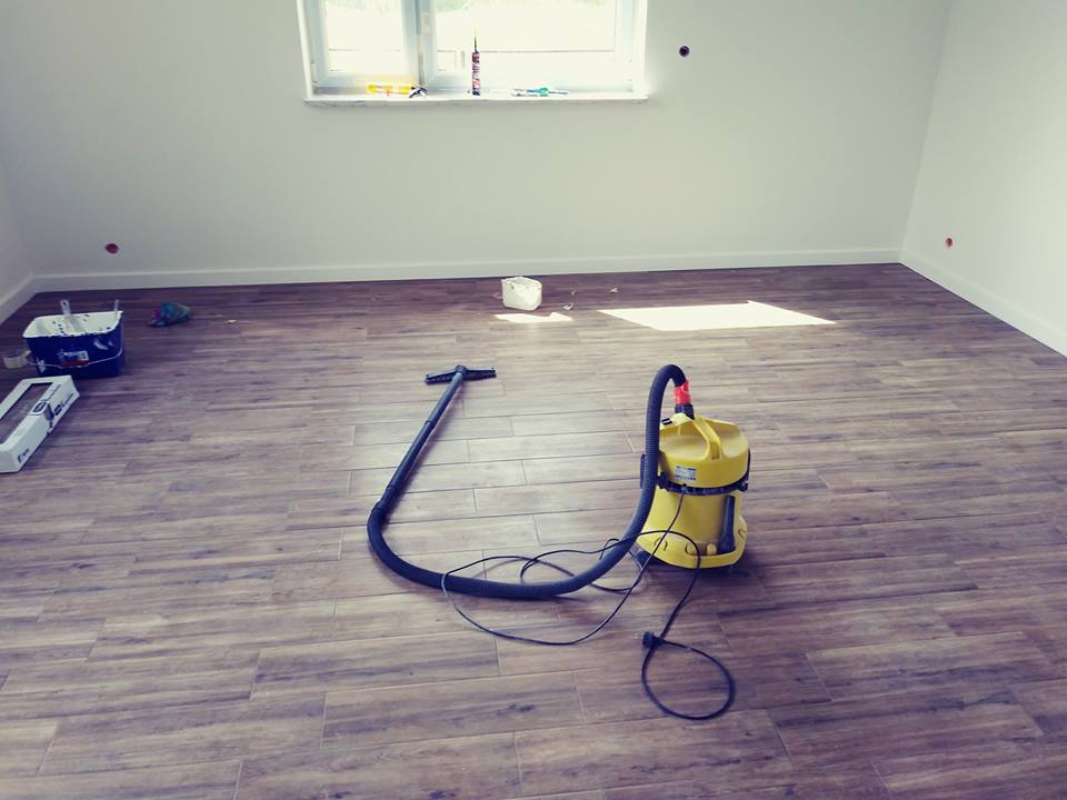 Pokój po ułożeniu podłogi z płytek imitujących drewno, widoczny odkurzacz przemysłowy, wiadro z farbą i akcesoria malarskie przy ścianie.