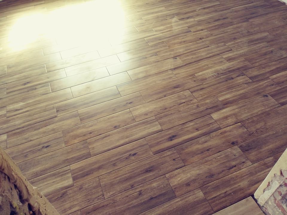 Ułożona pod kątem podłoga z płytek imitujących drewno, w ciepłych brązowych odcieniach, z widocznym fragmentem surowej ściany z cegły w rogu.