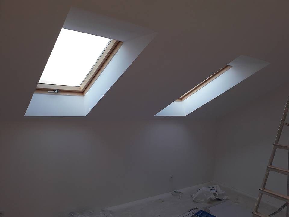 Poddasze po świeżym malowaniu na biało, dwa okna dachowe z drewnianą ramą, drabina oparta o ścianę, folia malarska na podłodze.
