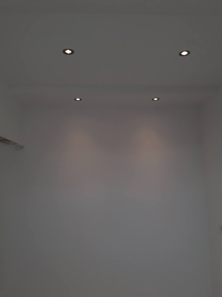 Biała ściana i sufit z czterema wbudowanymi, okrągłymi lampami halogenowymi, oświetlającymi ścianę. Widoczny fragment wentylacji sufitowej po lewej stronie.