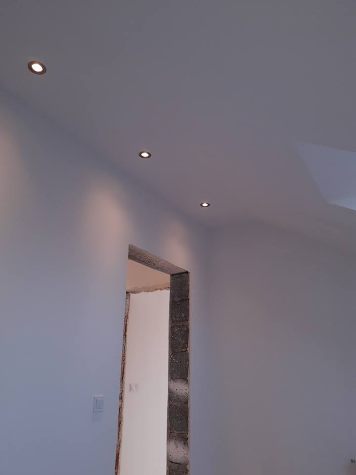 Świeżo pomalowane wnętrze z trzema punktowymi lampami sufitowymi i surowym otworem drzwiowym z widoczną cegłą.