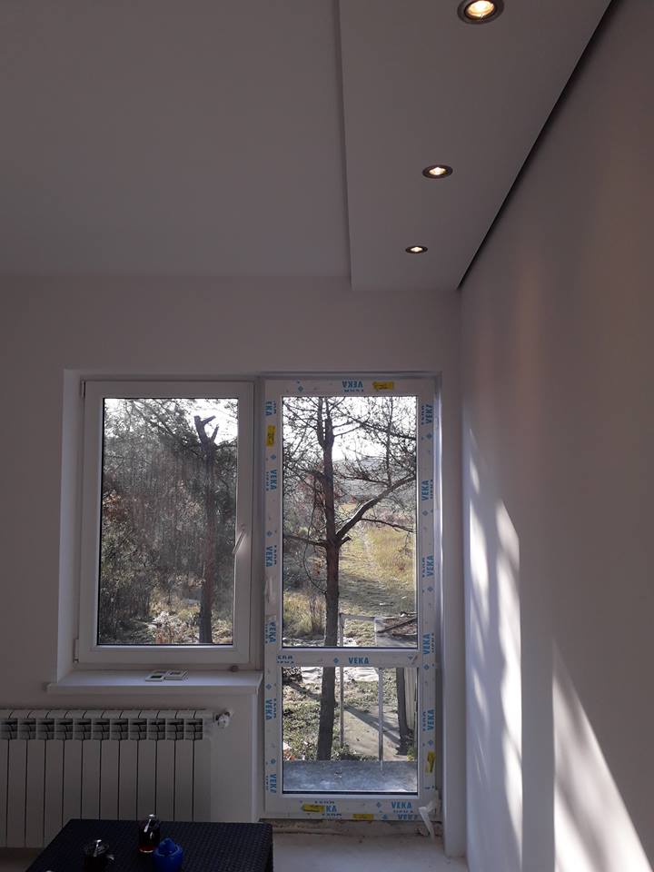 Nowe okna i drzwi balkonowe z białymi ramami VEKA w jasnym pokoju z grzejnikiem, widok na zewnątrz z drzewami.