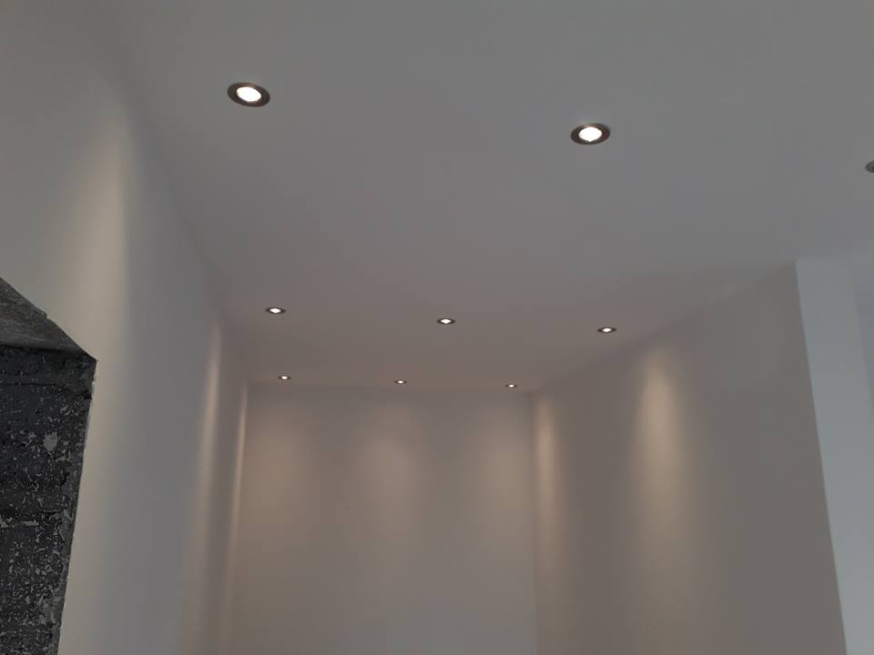 Wnętrze pomalowanego na biało pomieszczenia z zamontowanymi punktowymi lampami sufitowymi, widoczny fragment surowego betonowego elementu architektonicznego w rogu.