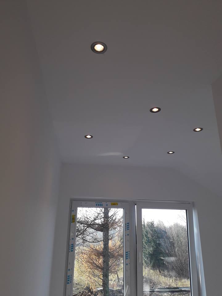 Białe wnętrze z oknem i sześcioma punktowymi lampami sufitowymi, widok na drzewa za oknem.