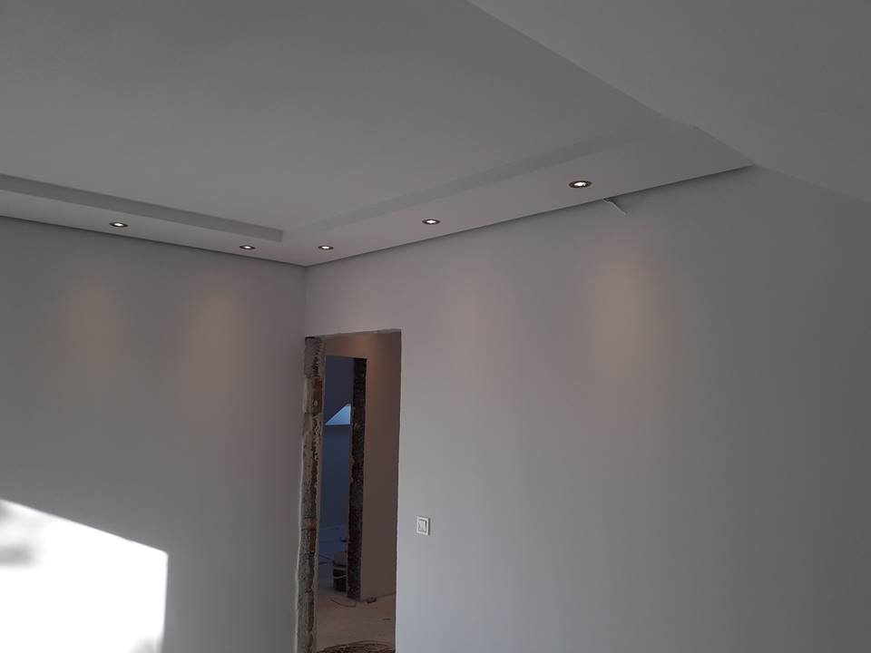 Wnętrze pokoju z pomalowanymi szarymi ścianami, widoczne oświetlenie punktowe w suficie podwieszanym i surowy otwór drzwiowy w trakcie prac wykończeniowych.