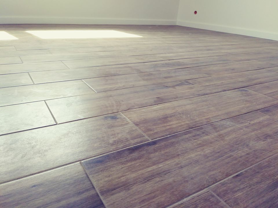 Ułożona podłoga z płytek imitujących drewno, z widocznymi fugami i fragmentem białej ściany z listwą przypodłogową oraz plamą światła słonecznego.