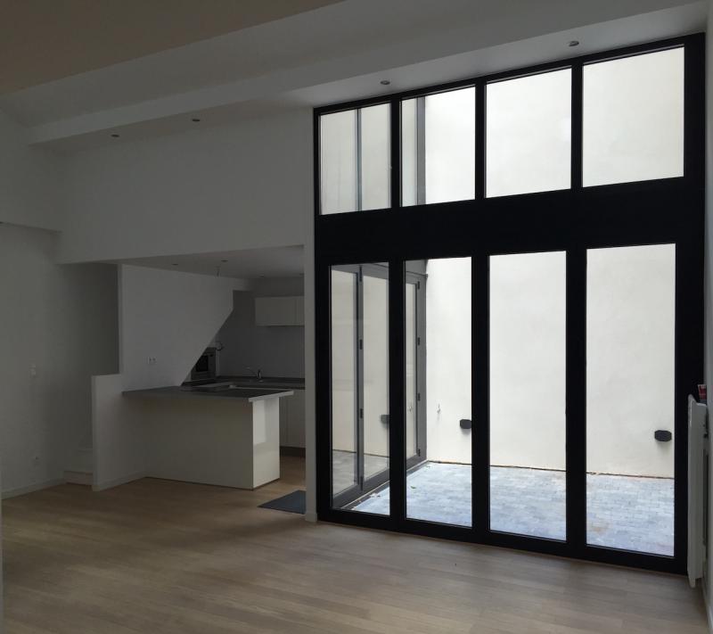 Wnętrze mieszkania z dużymi, przesuwnymi oknami z czarnymi ramami wychodzącymi na patio z szarą kostką brukową, widoczna drewniana podłoga i część aneksu kuchennego z blatem.