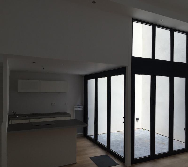 Wnętrze w trakcie wykańczania: surowa kuchnia z wyspą, minimalistyczne białe szafki wiszące, duże przesuwne okna z czarnymi ramami wychodzące na patio, drewniana podłoga, szara wycieraczka.