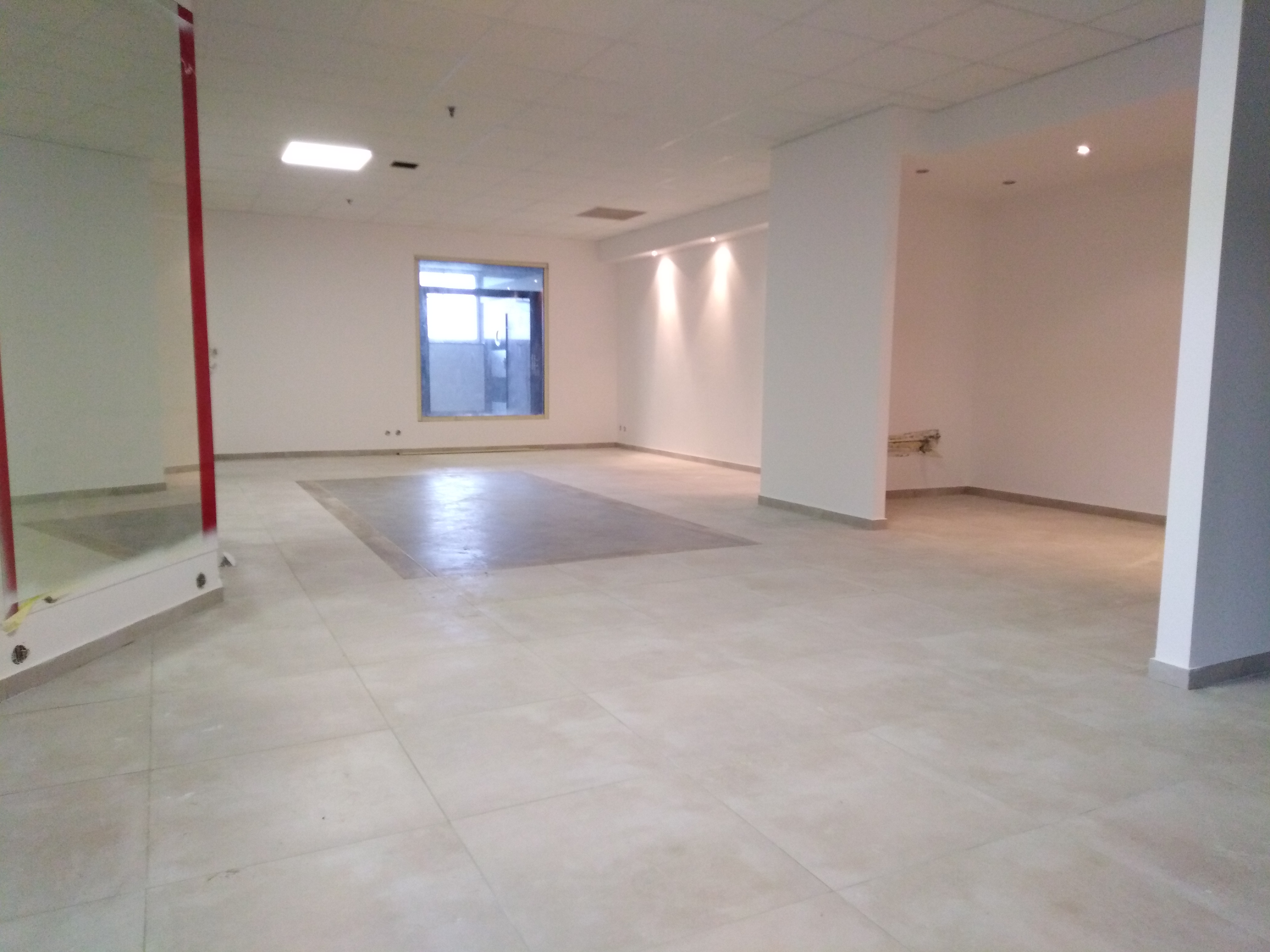 Przestronne, jasne wnętrze biurowe z beżową podłogą z płytek, białymi ścianami, kolumnami i oknem. W podłodze widoczny prostokątny obszar z innym odcieniem płytek. Na ścianie widoczne punkty świetlne.