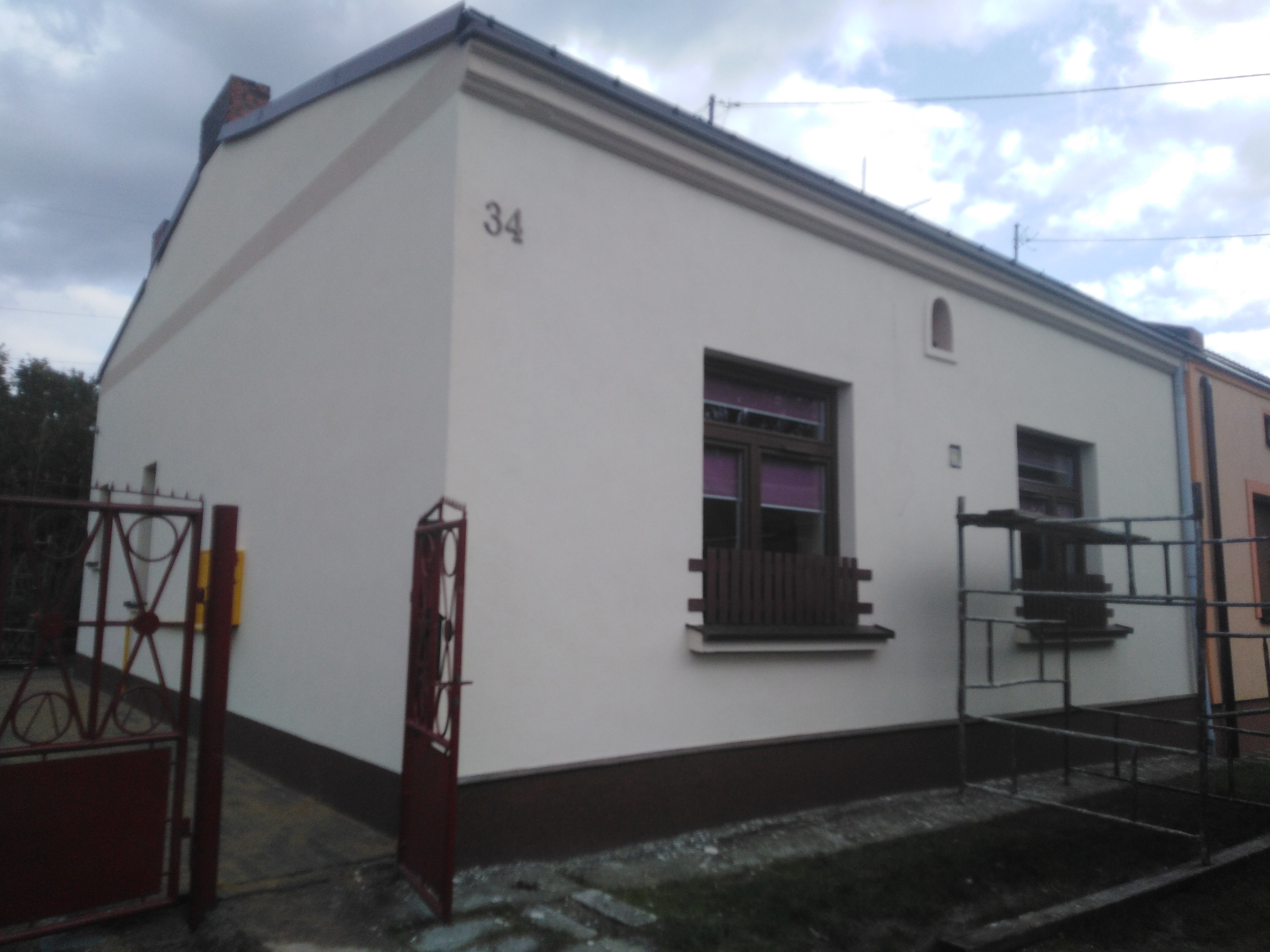 Odnowiona elewacja domu jednorodzinnego z numerem 34, widoczne rusztowanie przy ścianie bocznej, otwarte wejście na posesję.