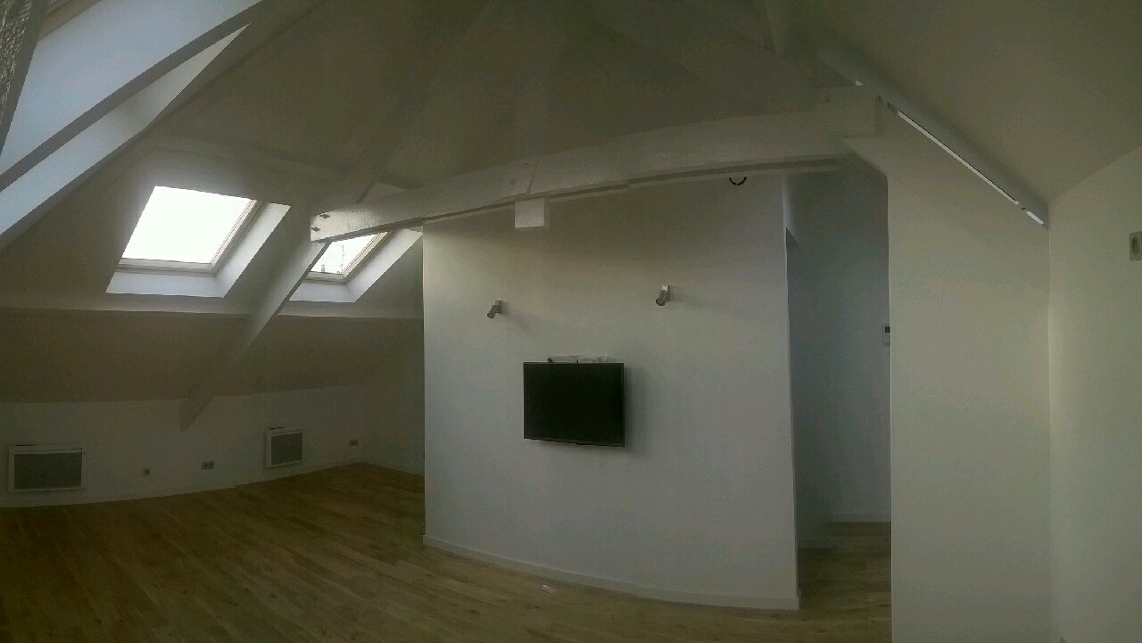 Jasne poddasze z drewnianą podłogą, skośnym sufitem z oknami dachowymi, białe ściany z zamontowanym telewizorem i punktowym oświetleniem.