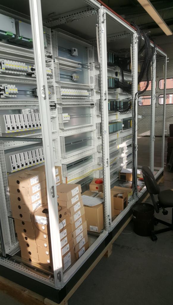 Rozdzielnica elektryczna w trakcie montażu, widoczne wewnętrzne komponenty, kable oraz stos kartonowych pudełek z elementami na palecie.