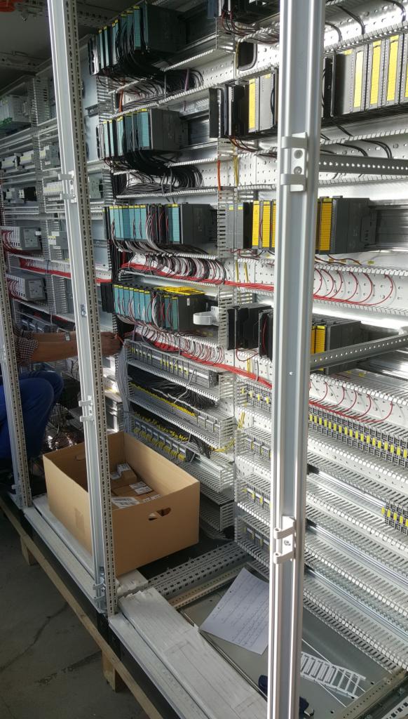 Rozbudowana szafa sterownicza w trakcie montażu, widoczne moduły PLC Siemens z okablowaniem, pracownik w tle oraz karton z elementami na podłodze.
