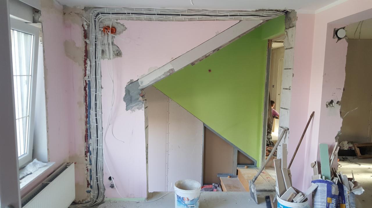 Wnętrze w trakcie remontu: widoczne przewody elektryczne prowadzone po ścianie, fragment konstrukcji schodów z zielonym elementem, ściany w kolorze pudrowego różu, narzędzia i materiały budowlane...