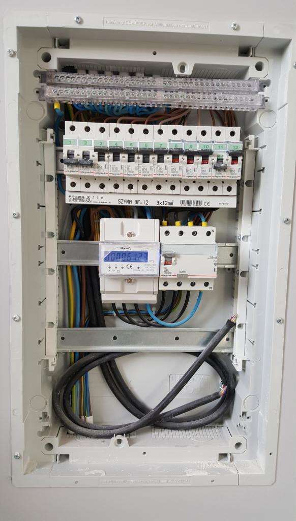 Wnętrze skrzynki elektrycznej z zamontowanym licznikiem energii, bezpiecznikami i okablowaniem w kolorach niebieskim, żółtym i brązowym.