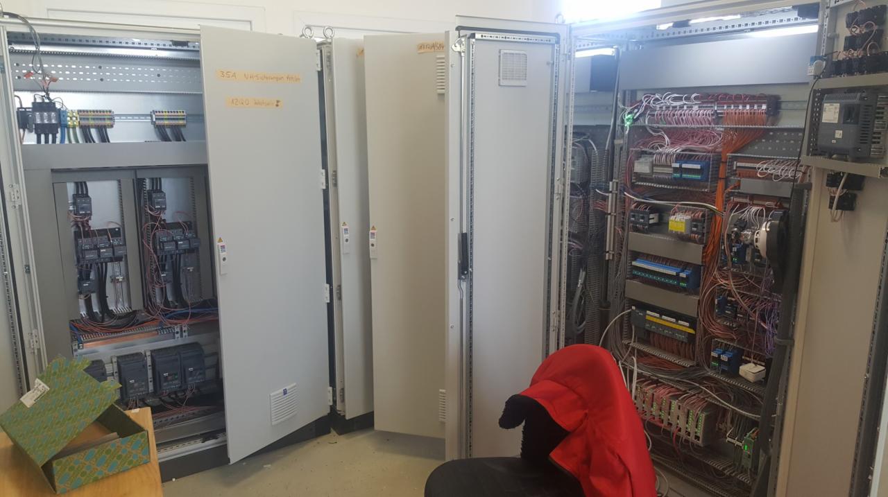 Otwarty panel sterowania z widocznymi przewodami i modułami, dodatkowo widoczne otwarte szafy sterownicze z żółtymi notatkami na drzwiach, otwarte pudełko na stole, a na krześle leży czerwona kurtka.