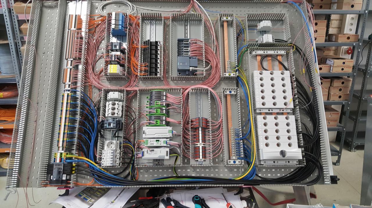 Rozbudowana szafa sterownicza w trakcie montażu, widoczne liczne przewody, moduły sterujące, listwy zaciskowe i elementy montażowe w metalowej obudowie, częściowo zakryta przez dokumentację...