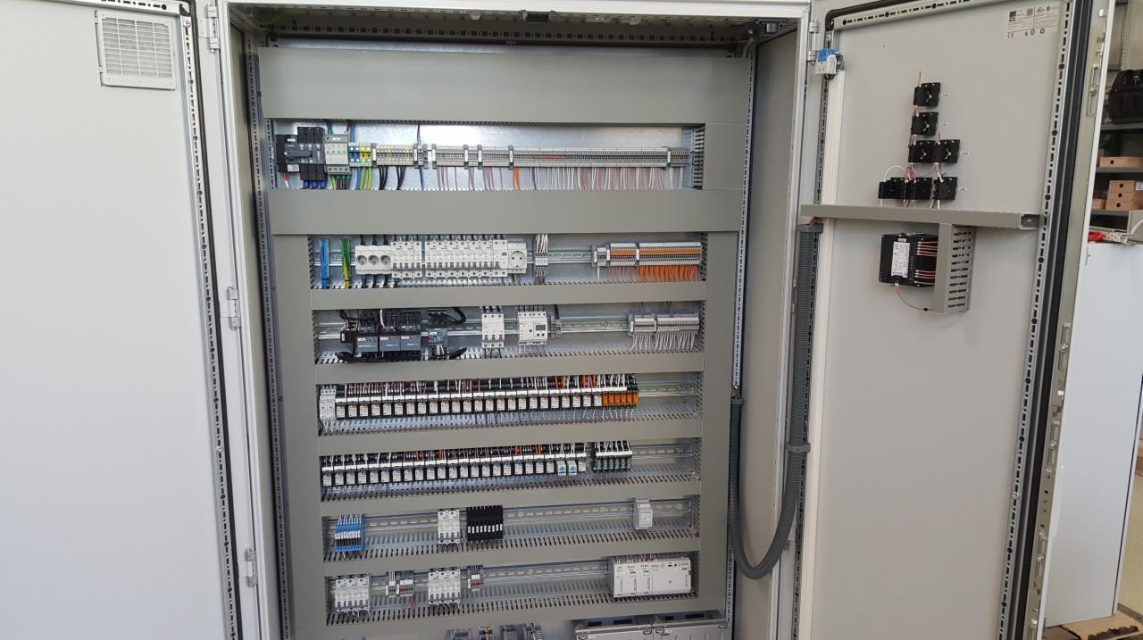 Otwarta szafa sterownicza z widocznymi elementami elektrycznymi, w tym liczne przekaźniki, bezpieczniki, listwy zaciskowe i okablowanie uporządkowane w poziomych rzędach.